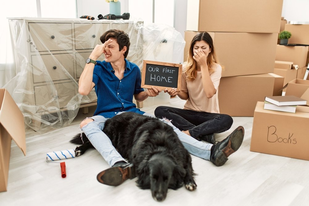 A young couple pinches their noses closed while sitting on the floor of their home in front of packed boxes while the dog lays in front of them smelling musty